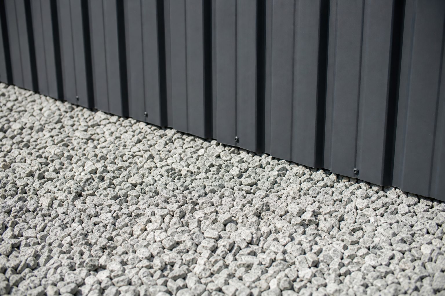Base of a dark Colorbond steel fence meeting clean grey gravel with no mulch or planting against the panel