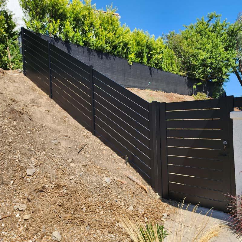 Horizontal aluminum slat fence and gate stepped down a graded hillside, with gaps between the slats